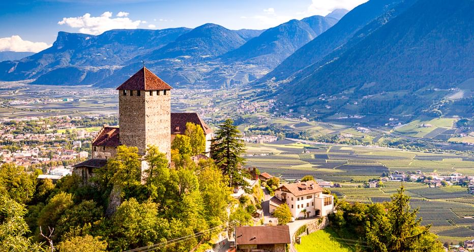 Mittelalterliche Burg mit Turm auf einem Hügel umgeben von grünen Bäumen, mit Blick auf das Etschtal und Berge in Südtirol unter blauem Himmel.