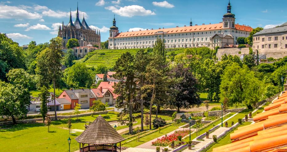 Panoramablick auf Kutná Hora mit gotischem Dom, barockem Schloss, terrassierten Gärten und Park mit Holzpavillon unter blauem Himmel.