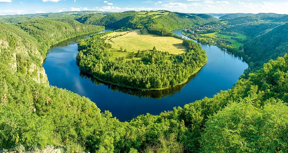 Luftaufnahme einer hufeisenförmigen Flussbiegung, umgeben von dichten Wäldern und grünen Wiesen unter blauem Himmel mit weißen Wolken in Tschechien.