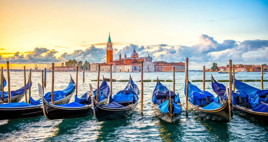 Reihe blau abgedeckter Gondeln an Holzpfählen in Venedig mit der Kirche San Giorgio Maggiore über der Lagune unter bewölktem Himmel bei Sonnenuntergang.
