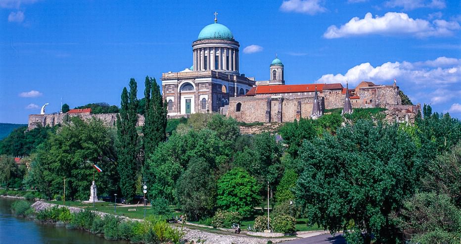 Die Basilika von Esztergom mit türkisfarbener Kuppel krönt einen Hügel über der Donau. Bäume säumen das Flussufer und ein Weg verläuft am Wasser.