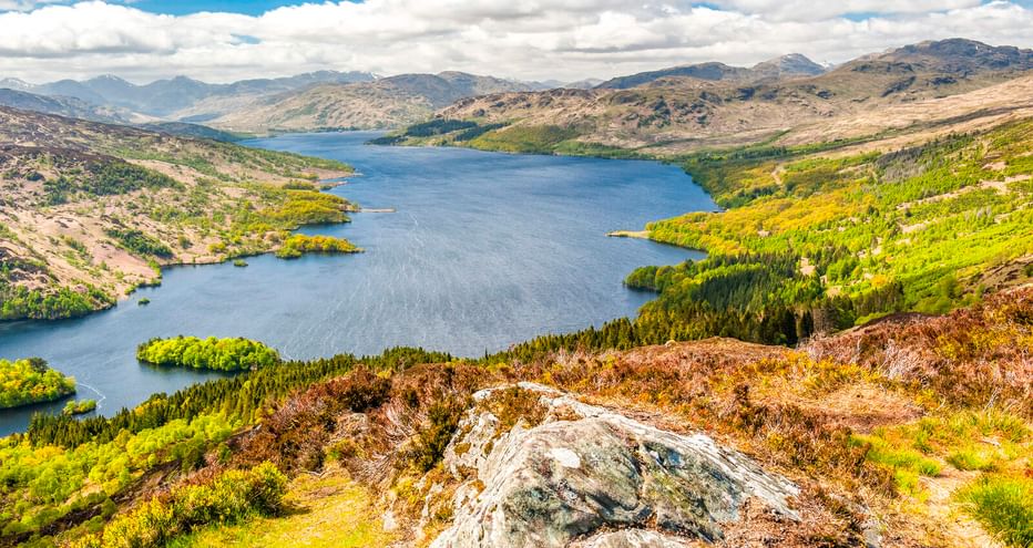 Panoramablick auf Loch Katrine, umgeben von grünen Wäldern und Hügeln. Felsiges Gelände im Vordergrund, blauer Himmel mit weißen Wolken.