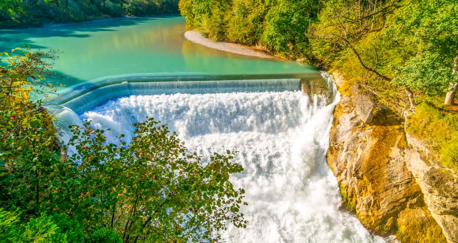 Luftaufnahme des Lechfalls bei Füssen mit türkisfarbenem Wasser, das über ein geschwungenes Wehr fließt, umgeben von dichtem grünem Wald und Felsen.