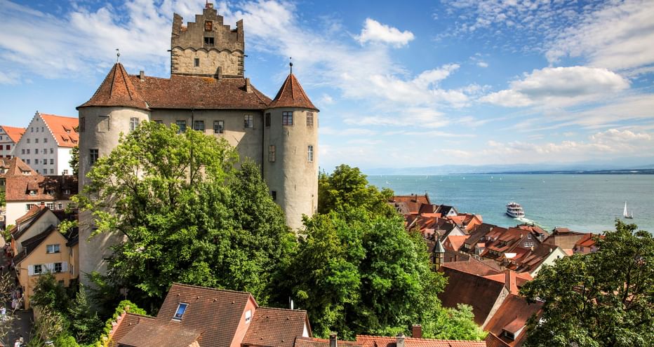 Burg Meersburg mit Turm und roten Ziegeldächern, umgeben von Bäumen, mit Blick auf den Bodensee mit Booten auf blauem Wasser unter bewölktem Himmel.