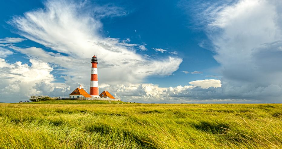 Rot-weiß gestreifter Leuchtturm Westerhever mit Nebengebäuden auf Westerheversand, umgeben von grünen Salzwiesen unter dramatischem Wolkenhimmel.