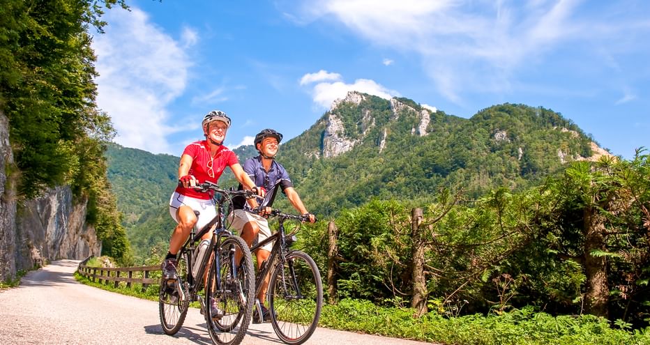 Zwei Radfahrer fahren nebeneinander auf einem asphaltierten Weg im oberösterreichischen Ennstal, mit grünen Bergen und blauem Himmel im Hintergrund.