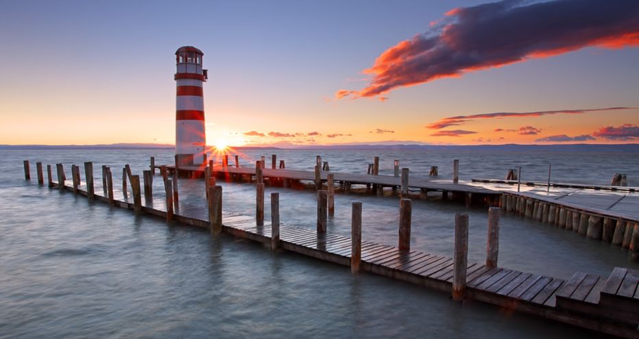Rot-weiß gestreifter Leuchtturm auf Holzsteg am Neusiedler See bei Sonnenuntergang mit orangefarbenen Wolken und ruhigem Wasser.