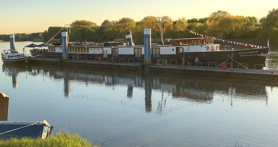 Flusskreuzfahrtschiff MS Magnifique III bei Sonnenuntergang mit bunten Flaggen vertäut. Bäume säumen das gegenüberliegende Ufer unter klarem Himmel.