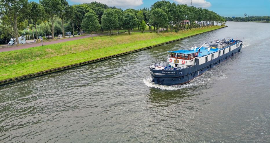 MS Magnifique III river cruise ship sailing on a Belgian canal lined with green trees. The blue and white vessel moves through calm water.