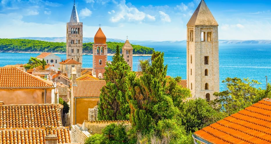 Blick auf die Altstadt von Rab mit vier Glockentürmen über Terrakottadächern. Das Adriatische Meer und grüne Küste im Hintergrund.