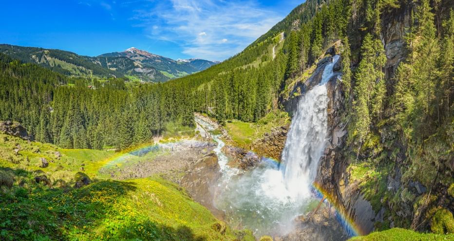 Krimmler Wasserfälle stürzen über Felsklippe mit Regenbogen im Sprühnebel, umgeben von grünen Almwiesen und dichtem Wald in Österreich.