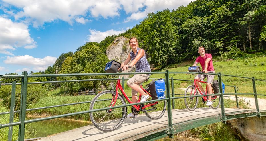 Zwei Radfahrer auf einer Metallbrücke über einen Bach bei Schelklingen. Grüne bewaldete Hügel und Felsformationen im Hintergrund unter blauem Himmel.