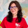 Krystsina mit dunklen, welligen Haaren und Brille trägt ein rotes Radweg-Reisen T-Shirt und lächelt vor einer bunten Weltkarte.