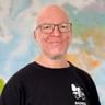 Portrait von Stefan G. mit Brille und schwarzem Radweg-Shirt, lächelnd vor einem bunten Weltkarten-Hintergrund.