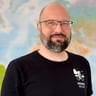 Porträt von Max mit Brille und schwarzem Radweg Reisen Shirt mit Fahrrad-Logo, stehend vor einer bunten Weltkarte.