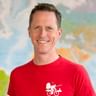 Thomas wearing a red cycling shirt with bicycle logo, smiling at camera. World map visible in blurred background.