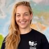 Anja mit langen blonden Haaren lächelnd, trägt ein schwarzes Radweg-Shirt mit Radfahrer-Logo. Im Hintergrund ist eine bunte Weltkarte sichtbar.