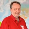 Porträt von Claus in einem roten Poloshirt mit Fahrrad-Logo, stehend vor einer bunten Weltkarte im Hintergrund.