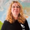Porträt von Adriana mit Brille und welligem blondem Haar, trägt ein schwarzes Shirt mit Fahrrad-Logo, steht vor einer Weltkarte.