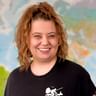 Yana mit lockigem braunem Haar und gelben Ohrringen, breit lächelnd in einem schwarzen Fahrrad-T-Shirt. Im Hintergrund ist eine bunte Weltkarte sichtbar.
