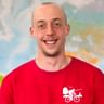 Porträt von Tim S in einem roten Shirt mit Fahrrad-Logo, lächelnd in die Kamera. Im Hintergrund ist eine bunte Karte sichtbar.