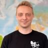 Portrait von Raul in schwarzem T-Shirt mit Fahrrad-Logo, lächelnd vor einer Weltkarte im Hintergrund.