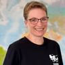 Beate mit Brille und schwarzem Shirt mit Fahrrad-Logo, lächelnd vor einer bunten Weltkarte.