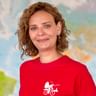 Ilka mit lockigem braunen Haar trägt ein rotes Shirt mit Fahrrad-Logo und lächelt vor einem unscharfen Weltkartenhintergrund.