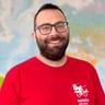 Portrait von Tim J. mit Brille und rotem Radweg Reisen Shirt, lächelnd vor einem Weltkarten-Hintergrund.