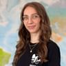 Porträt von Nina mit Brille und langen welligen braunen Haaren, trägt ein schwarzes Shirt mit Radfahrer-Grafik. Eine Weltkarte ist im Hintergrund sichtbar.