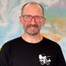 Porträt von Lars mit Brille und schwarzem Shirt mit Fahrrad-Logo, lächelnd vor einer Weltkarte im Hintergrund.
