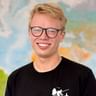 Peter mit blonden Haaren und schwarzer Brille, der breit lächelt. Er trägt ein schwarzes T-Shirt. Im Hintergrund ist eine bunte Karte sichtbar.