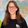 Annika mit Brille und langen braunen Haaren, lächelnd vor einer Weltkarte. Sie trägt ein schwarzes Shirt mit Fahrrad-Logo.