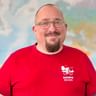 Porträt von Alex mit Brille und rotem Radweg Reisen T-Shirt mit Firmenlogo, lächelnd vor einer Weltkarte im Hintergrund.