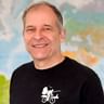 Portrait von Christoph mit kurzem grauen Haar, der breit lächelt. Er trägt ein schwarzes Shirt mit weißer Fahrradgrafik. Eine bunte Karte ist hinter ihm sichtbar.